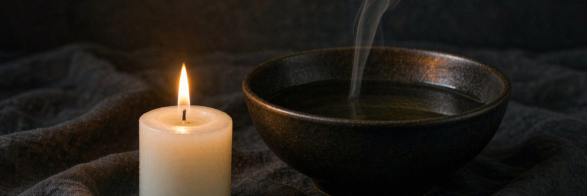 A white candle and a bowl of still water with curling smoke on dark linen in amber light